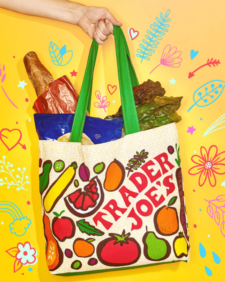 Someone holding a trader joe's bag full of groceries in front of a yellow background with illustrations of flowers, hearts, rainbows