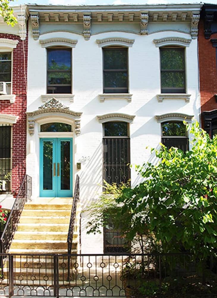 Two-story townhouse with white facade, turquoise door, and ornate trim, flanked by red brick buildings and greenery.