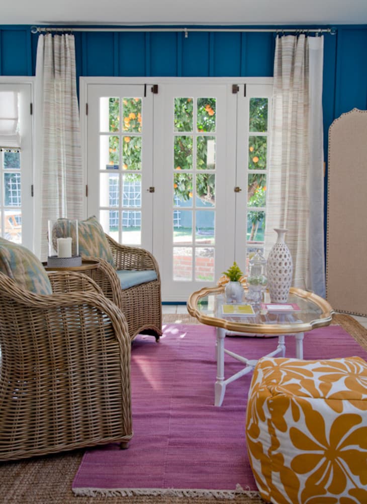 Wicker chairs and a glass coffee table on a purple rug in a sunroom with blue walls and French doors.