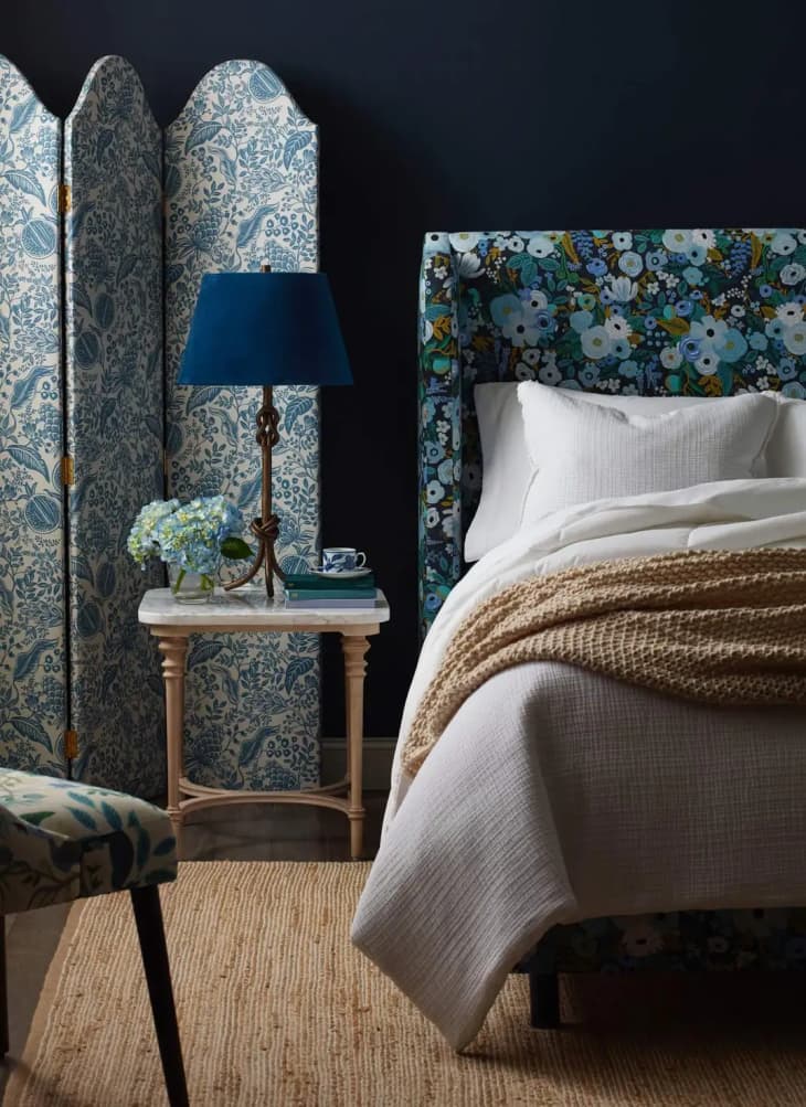 Bedroom with floral upholstered bed, blue lamp, and floral room divider. Beige knit throw on white bedding.