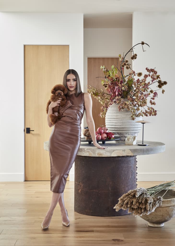 Woman in brown dress holding a small dog, standing by a round table with a large vase of dried flowers and pomegranates.