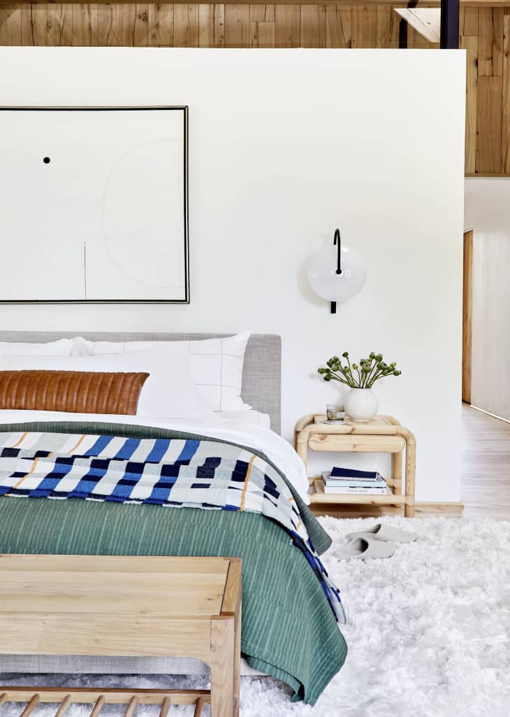 Modern bedroom with a wooden bench, abstract art, and a cozy bed with layered blankets and pillows.