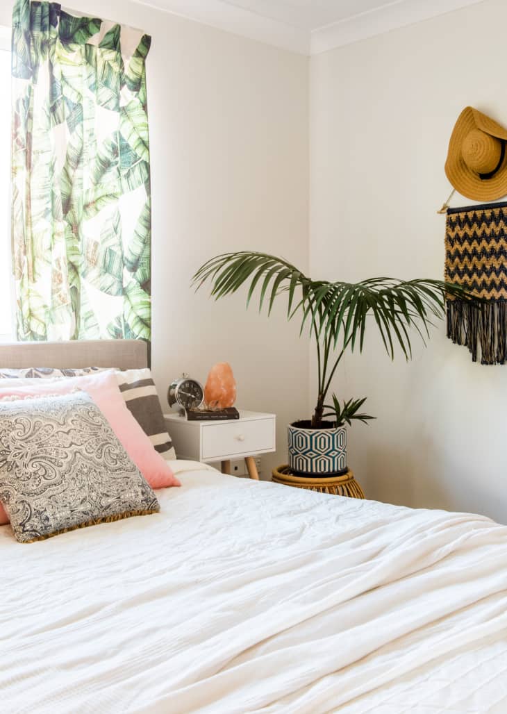 Bedroom with a white bed, patterned pillows, leafy curtains, a potted plant, and a straw hat on the wall.