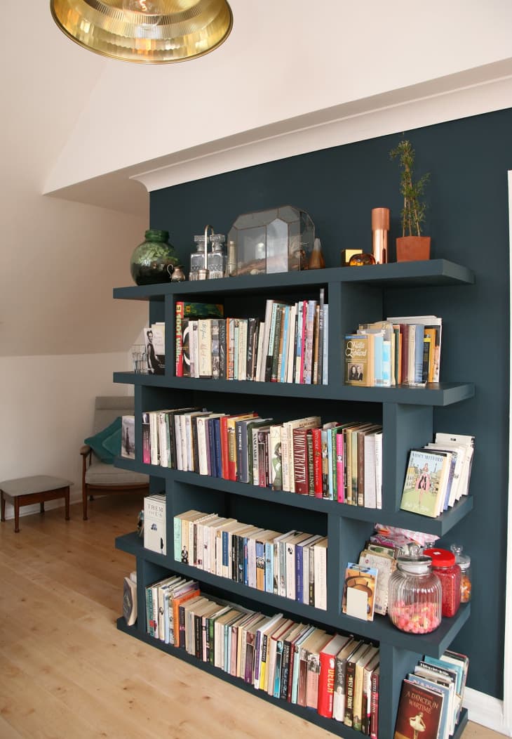 Dark blue bookshelf filled with books, glass jars, and decor items, set against a wooden floor and white wall.