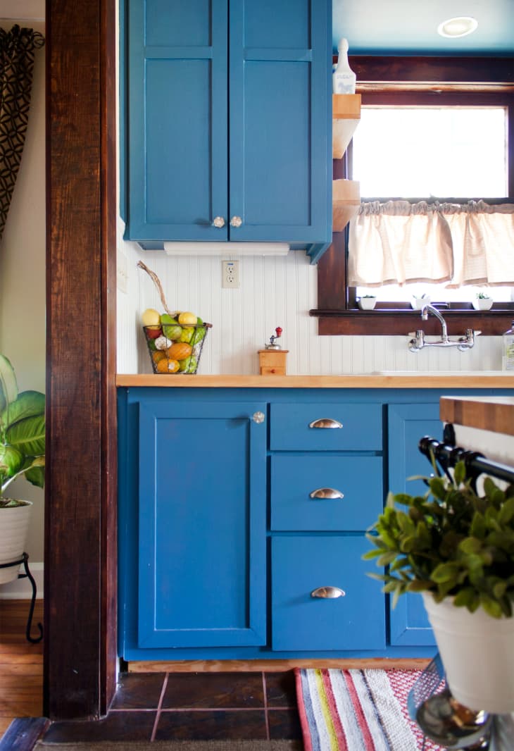 Blue kitchen cabinets with wooden countertop, fruit basket, and striped rug near a window with beige curtains.