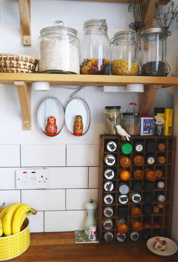 Kitchen shelf with glass jars of grains and pasta, spice rack, bananas, and Russian nesting dolls on a tiled wall.