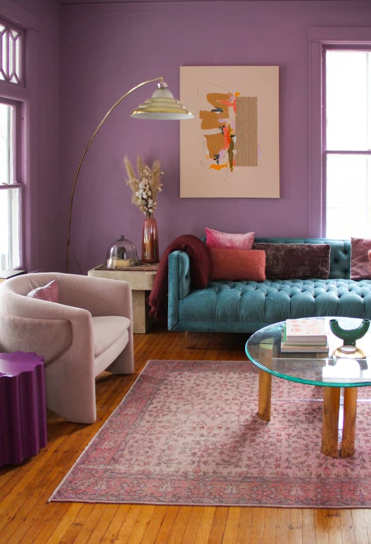 Living room with purple walls, teal sofa, abstract art, pink armchair, glass coffee table, and floral rug.