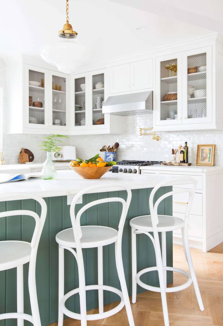 White kitchen with green island, white bar stools, glass cabinets, and a bowl of oranges on the counter.