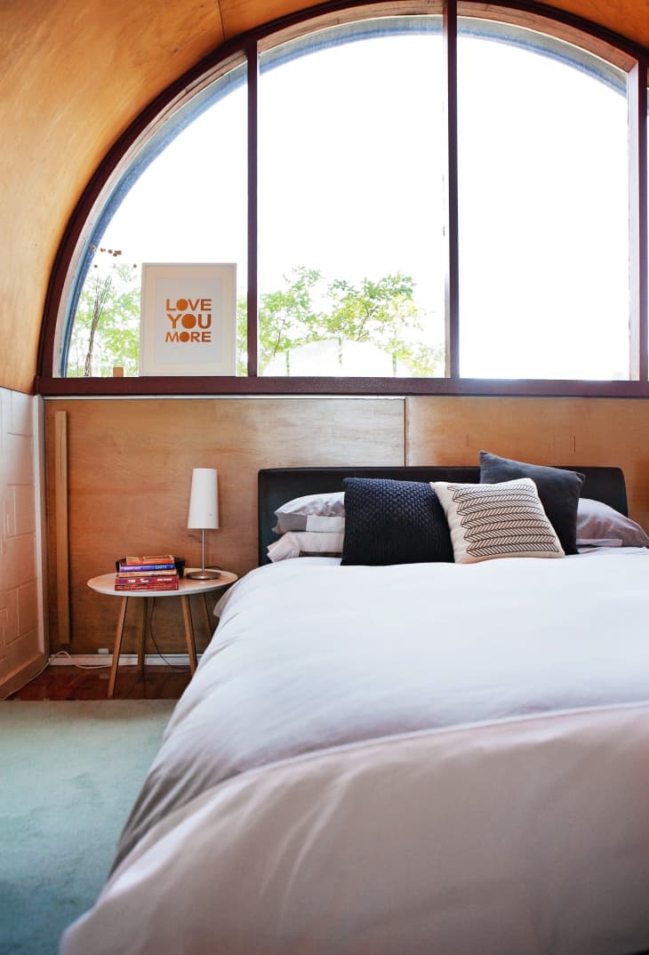 Cozy bedroom with arched window, white bedding, side table with books, lamp, and "Love You More" sign.