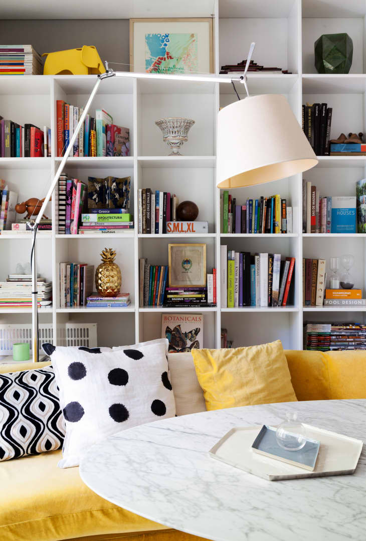 Bookshelf with colorful books, modern lamp, yellow sofa, and patterned cushions in a cozy living room.