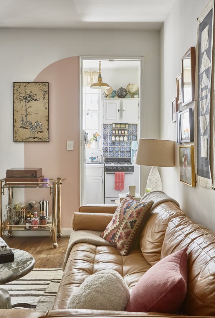 Living room with tan leather sofa, colorful pillows, bar cart, and view into kitchen with patterned tiles and globe decor.