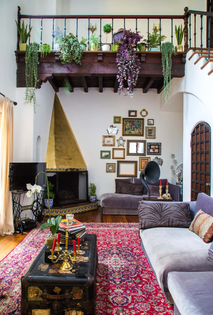 Living room with vintage decor, red patterned rug, gray sofas, gold fireplace, and plants on a wooden balcony.