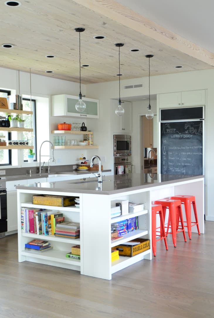 Modern kitchen with a large island, open shelves with books, red stools, pendant lights, and a chalkboard wall.