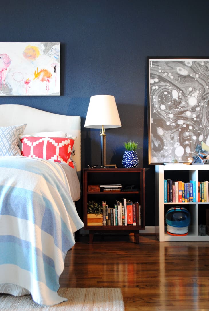 Bedroom with navy walls, abstract art, a bed with colorful pillows, a nightstand with books, and a lamp.