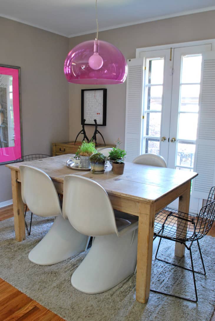 Dining room with wooden table, white and black chairs, pink pendant light, and potted plants on a beige rug.