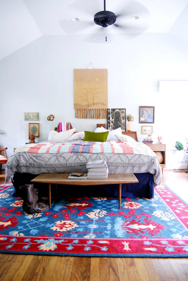 Bedroom with a colorful quilted bed, blue and red patterned rug, wooden bench, and eclectic wall art.
