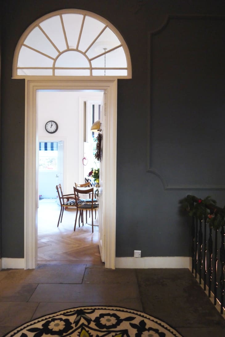 View through arched doorway into dining room with wooden chairs, table, wall clock, and floral arrangement.