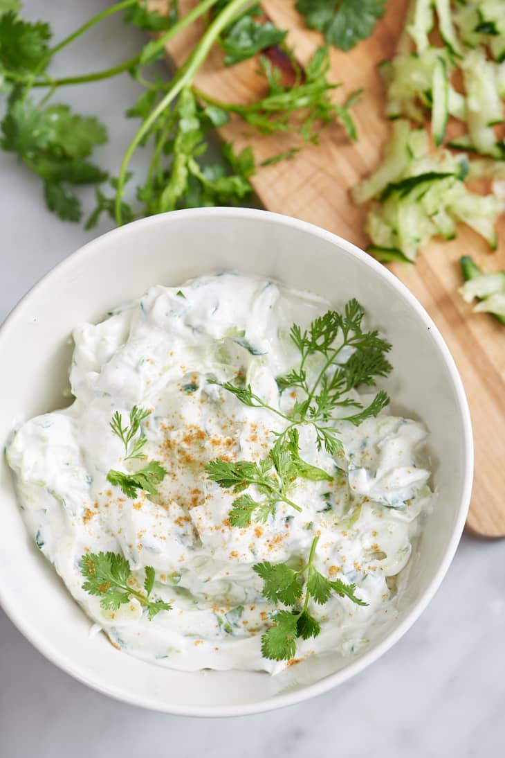 Creamy raita garnished with fresh cilantro in a white bowl, next to chopped cucumber on a wooden board.