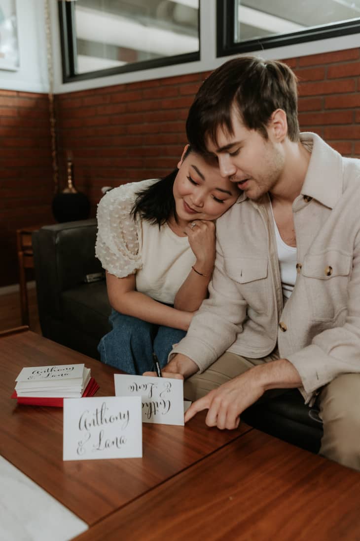 Couple sitting on a sofa, writing wedding invitations labeled "Anthony & Lana" on a wooden table.