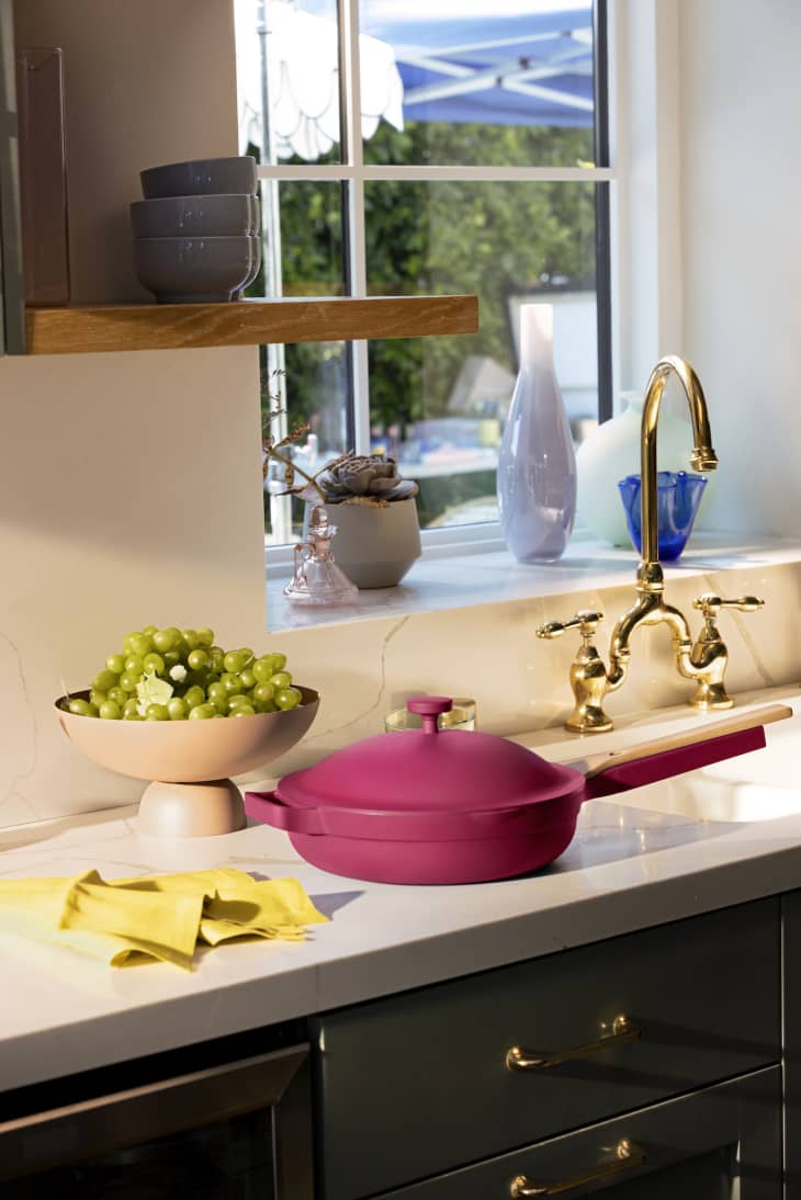 Pink pan on kitchen counter with green grapes in a bowl, yellow cloth, and gold faucet near window.