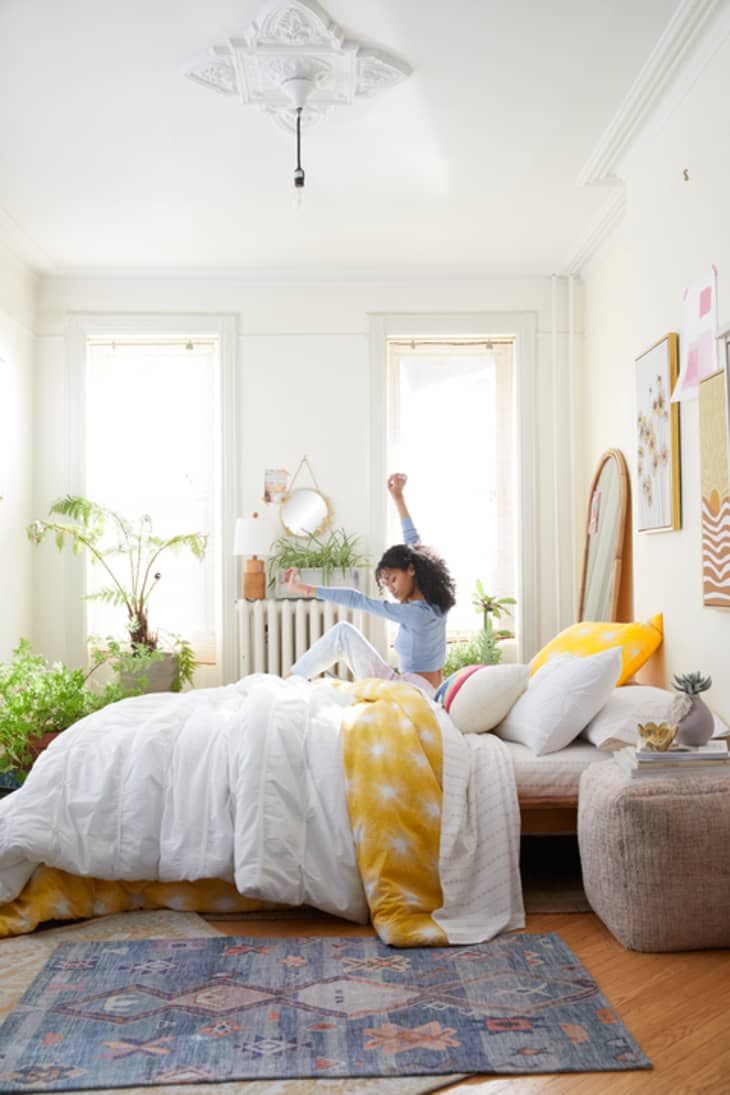Woman stretching on a bed with white and yellow bedding, surrounded by plants and wall art in a bright bedroom.