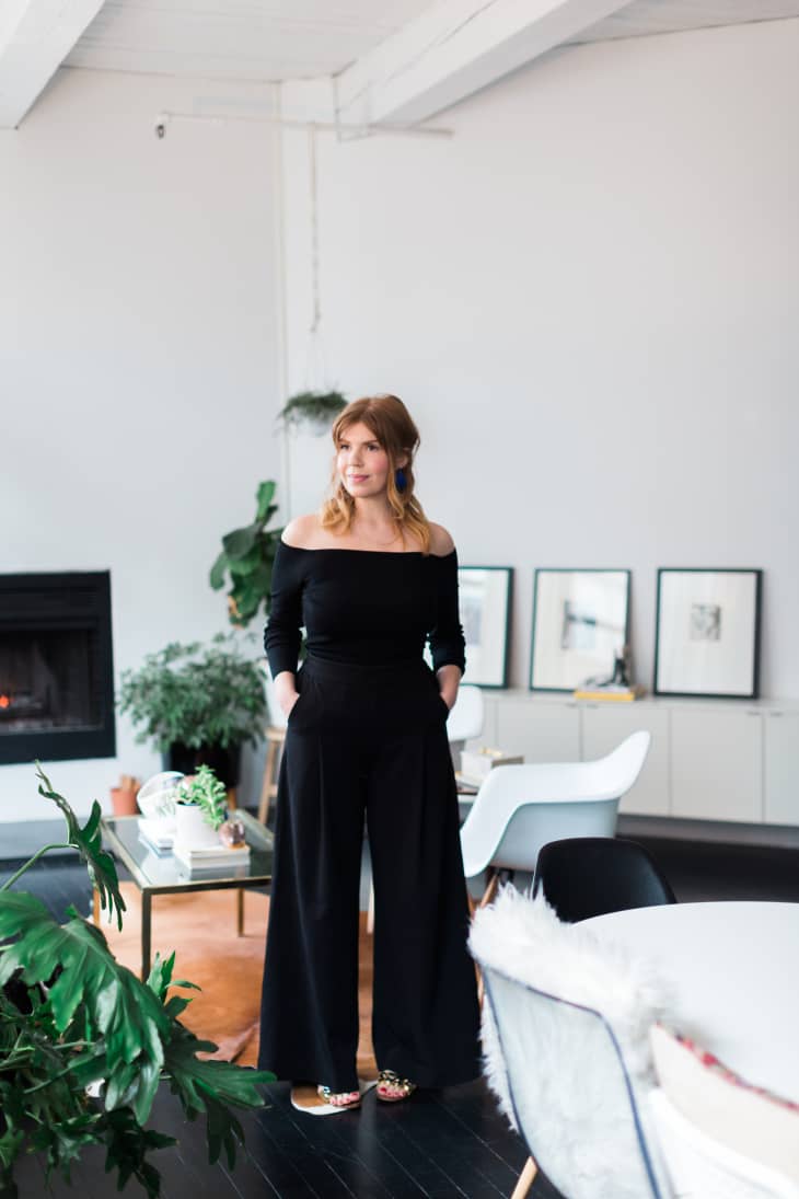 A woman wearing a black off-shoulder top and wide-legged pants poses in her living space