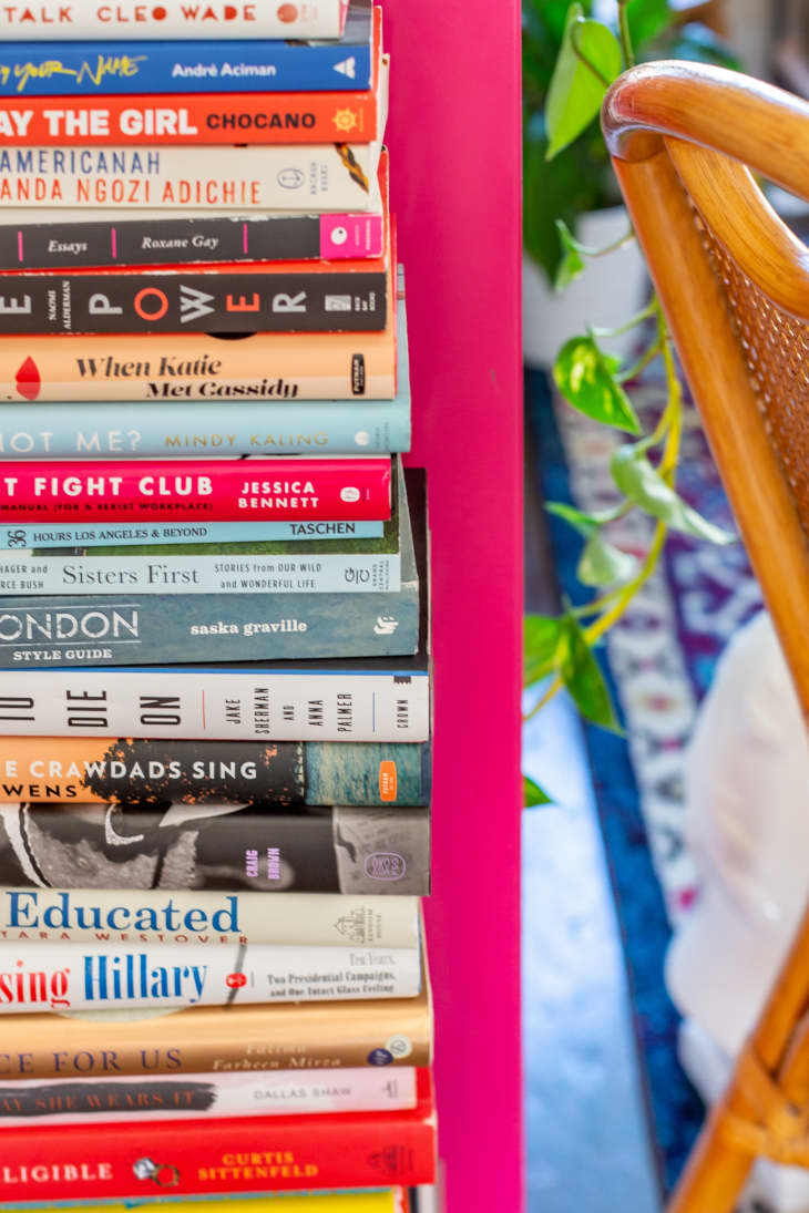 Stack of colorful books on a pink shelf next to a wooden chair and green plant.