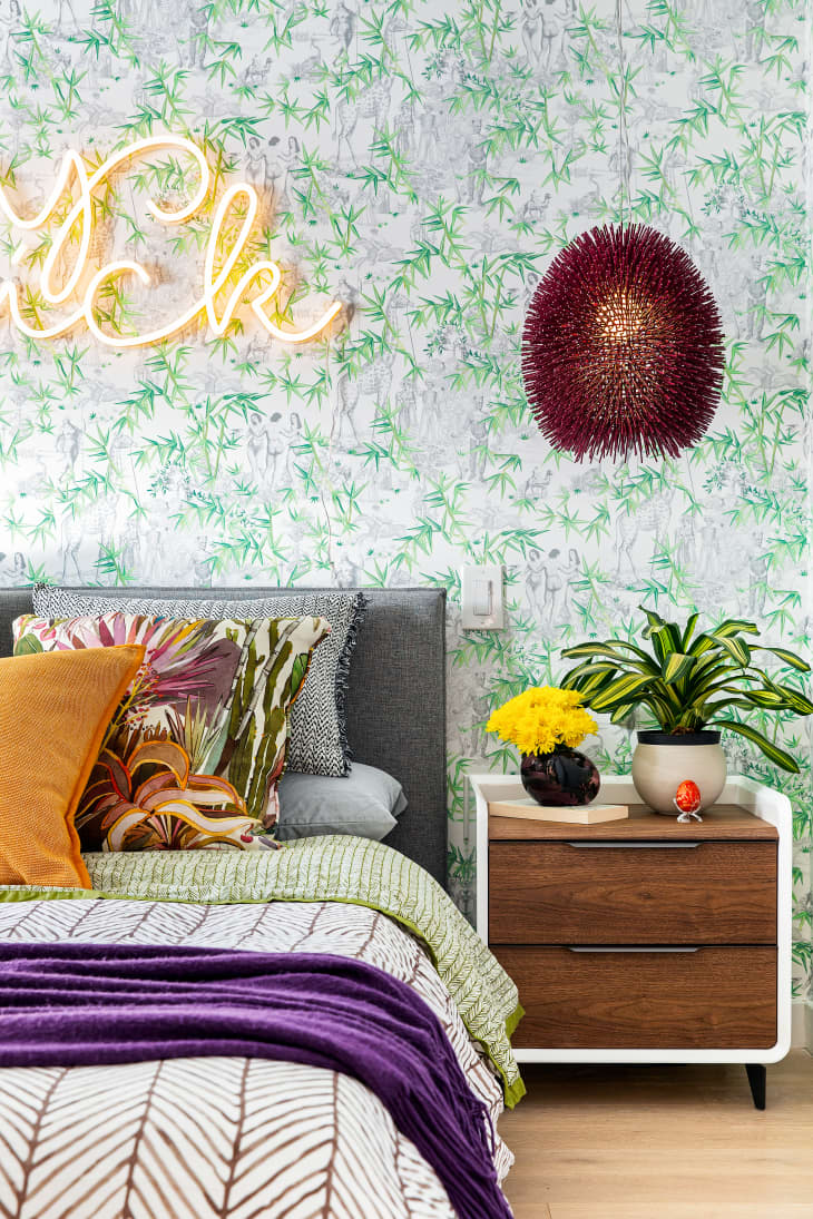 Bedroom with floral wallpaper, neon sign, colorful pillows, wooden nightstand, yellow flowers, and a potted plant.
