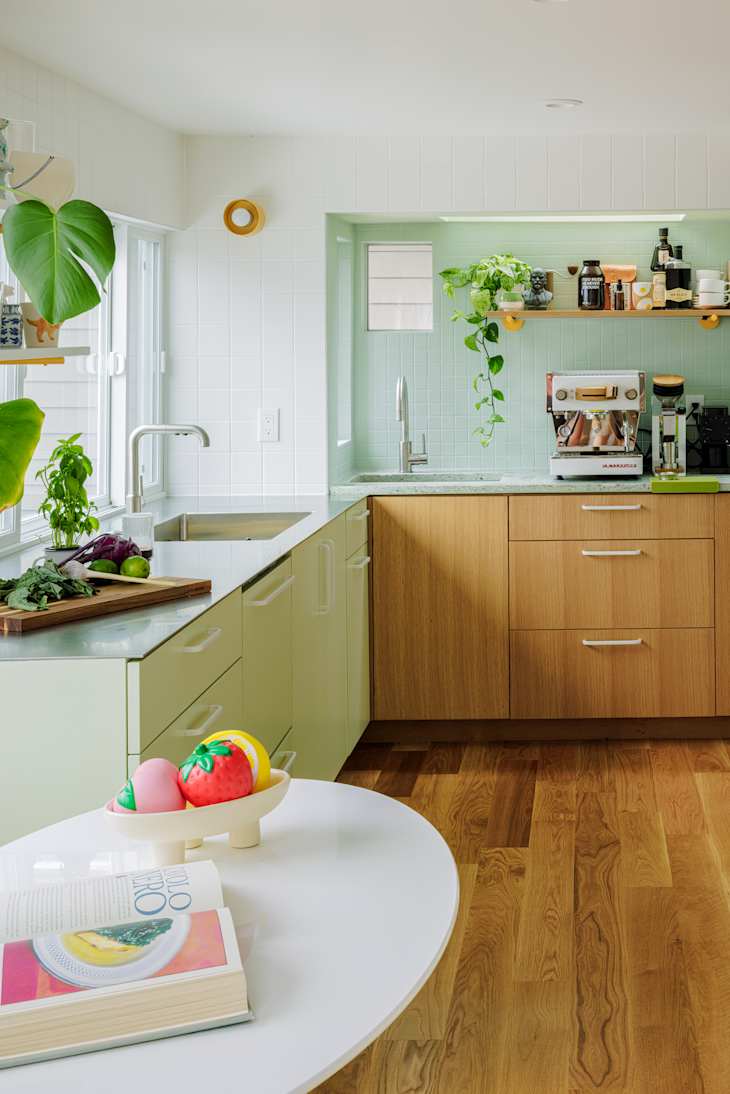 Modern kitchen with light green cabinets, wooden drawers, a white round table, and colorful decorative fruit.