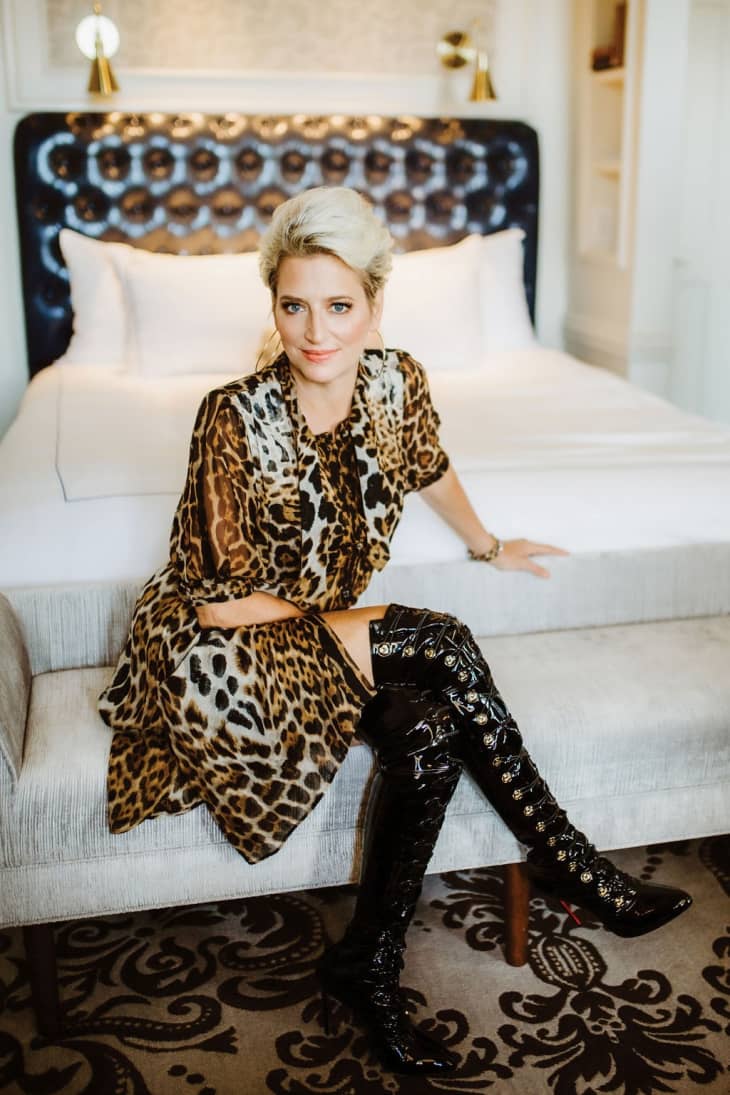Woman in leopard print dress and black knee-high boots sitting on a bench in a stylish bedroom.