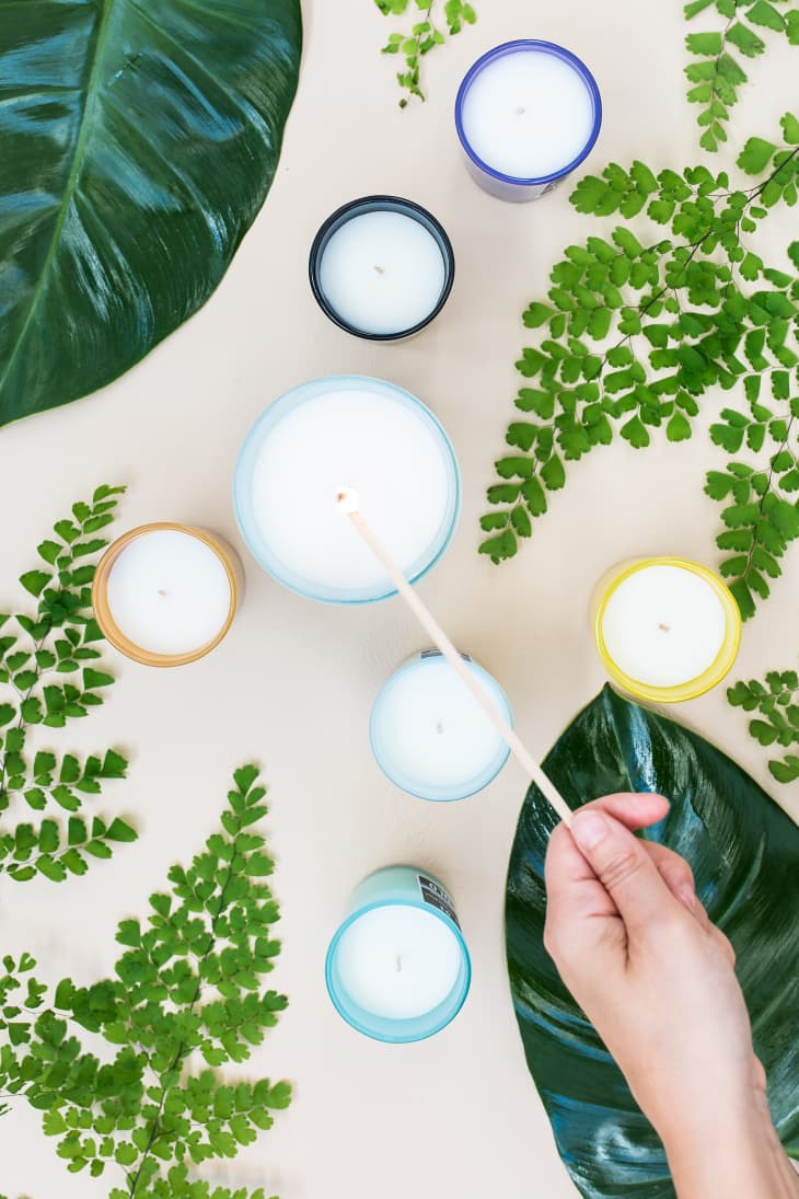 Hand lighting candles surrounded by green leaves on a beige surface.