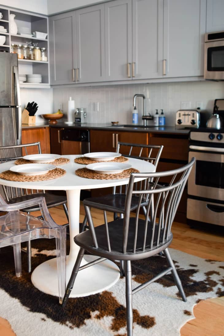 Modern kitchen with gray cabinets, round white table, black chairs, and cowhide rug. Stainless steel appliances and open shelving.