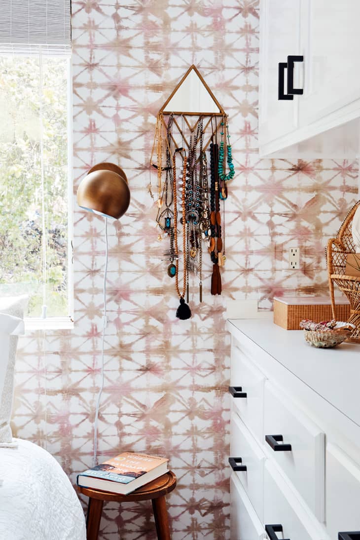 Bedroom corner with patterned wallpaper, jewelry hanging on a wall organizer, white dresser, and a small wooden stool with books.