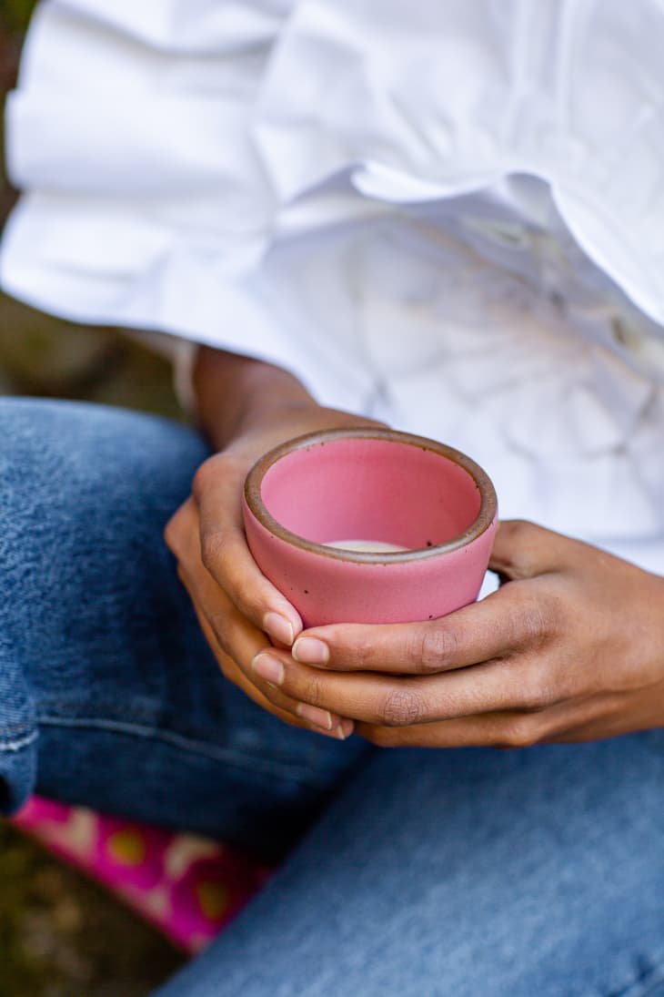Person holding a pink ceramic cup while sitting, wearing blue jeans and a white shirt.