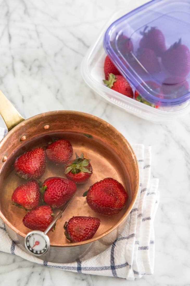 Strawberries in a copper pot with a thermometer, next to a container of strawberries on a marble surface.