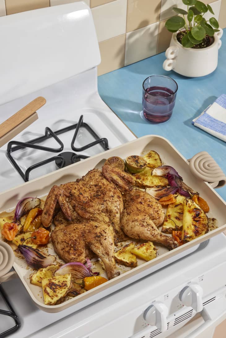 Roasted spatchcock chicken with pineapple, red onion, and peppers on a baking tray atop a stove, next to a plant and glass.