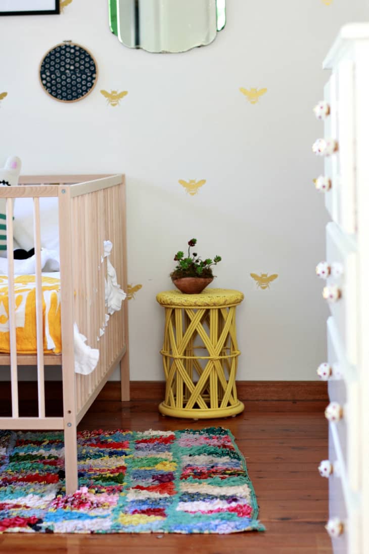 Nursery with wooden crib, colorful patchwork rug, yellow side table, and potted plant against a wall with bee decals.
