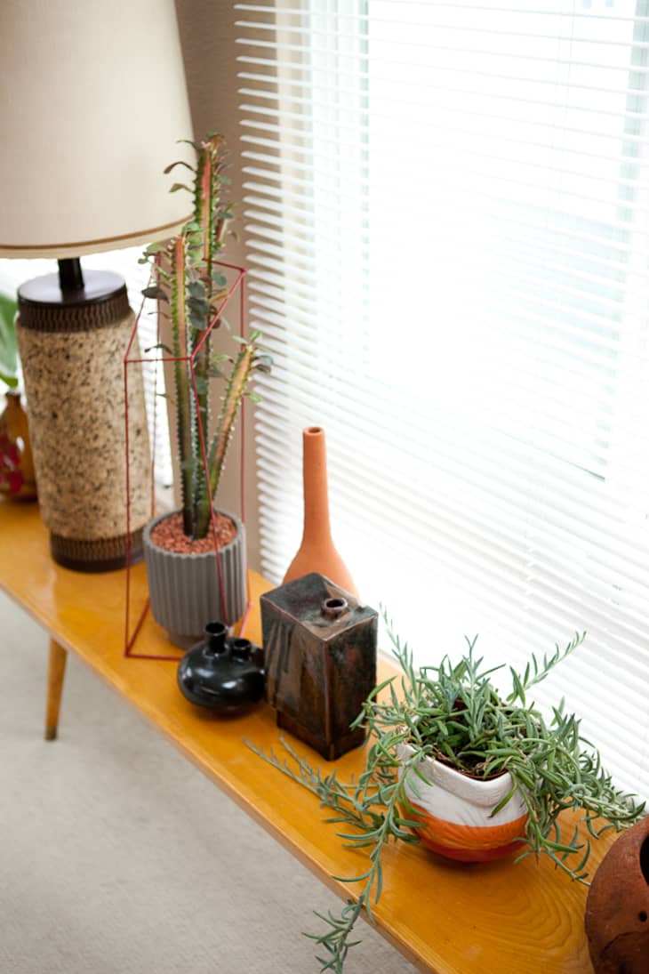 Wooden console table with potted plants, ceramic vases, and a lamp near window blinds.