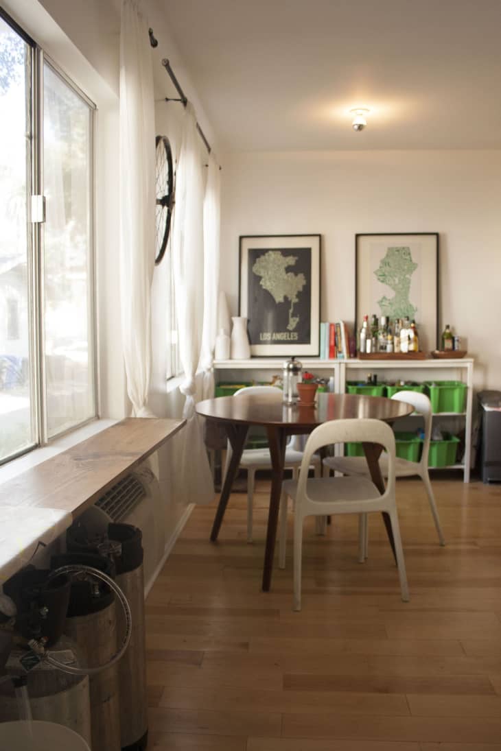 Dining area with round wooden table, white chairs, wall art, and a shelf with books and bottles.