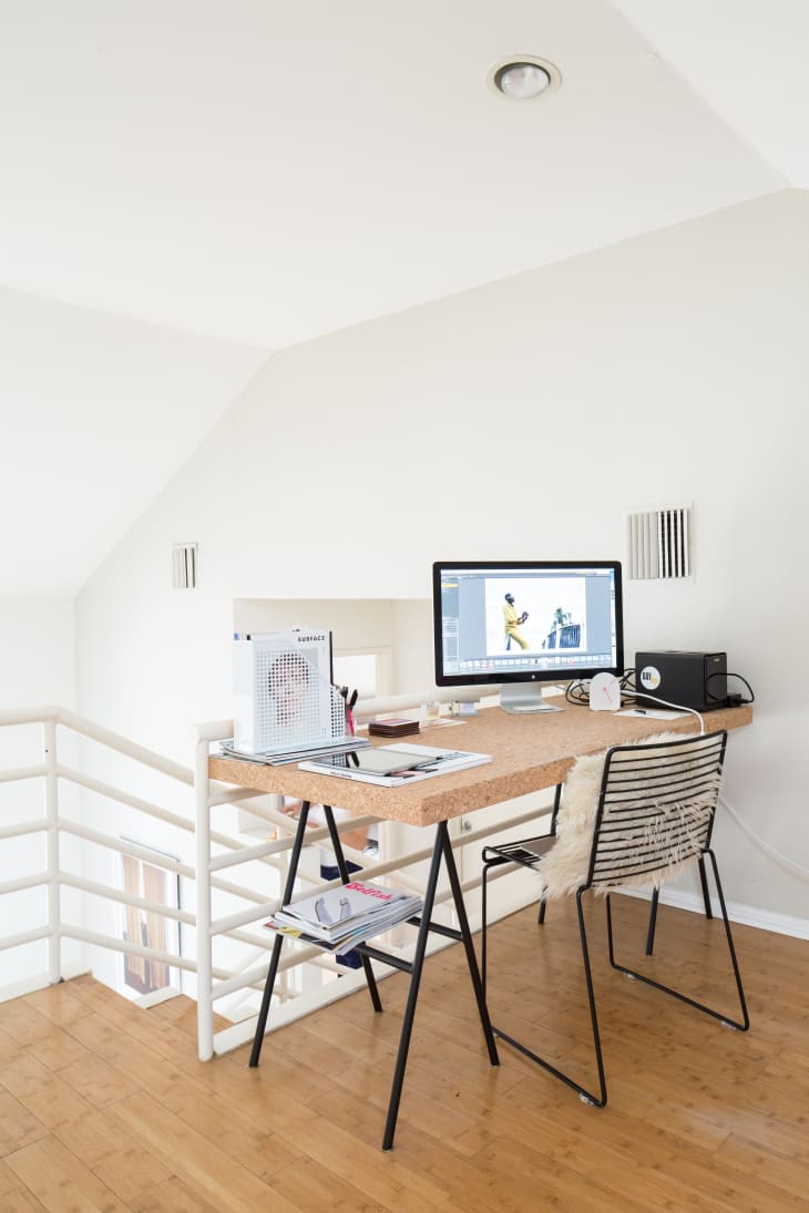 Home office with a wooden desk, computer monitor, wire chair, and magazines on a light wood floor.