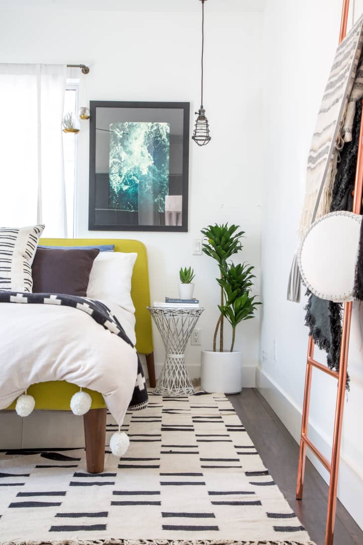Modern bedroom with a yellow bed, black and white decor, abstract art, and a potted plant on a geometric side table.