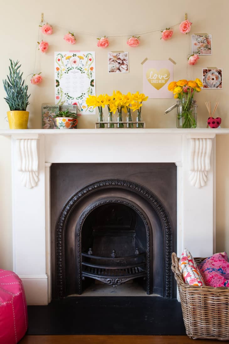 White fireplace with black insert, daffodils in vases, pink roses garland, and decorative prints on the wall.