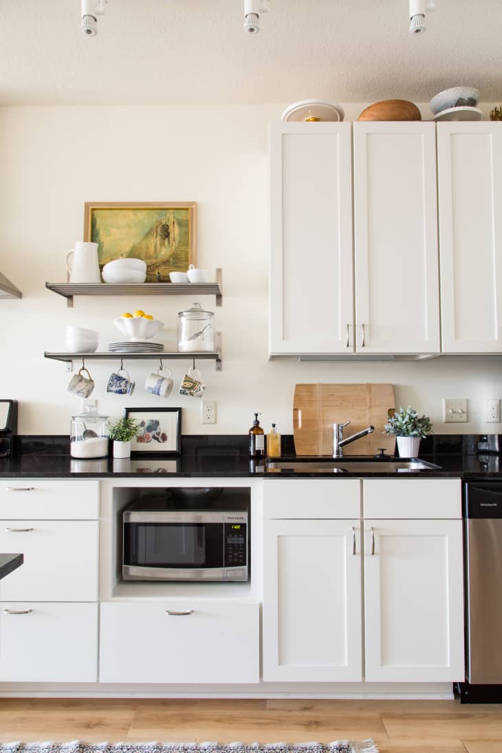 Modern kitchen with white cabinets, black countertop, microwave, open shelves with decor, and potted plants.