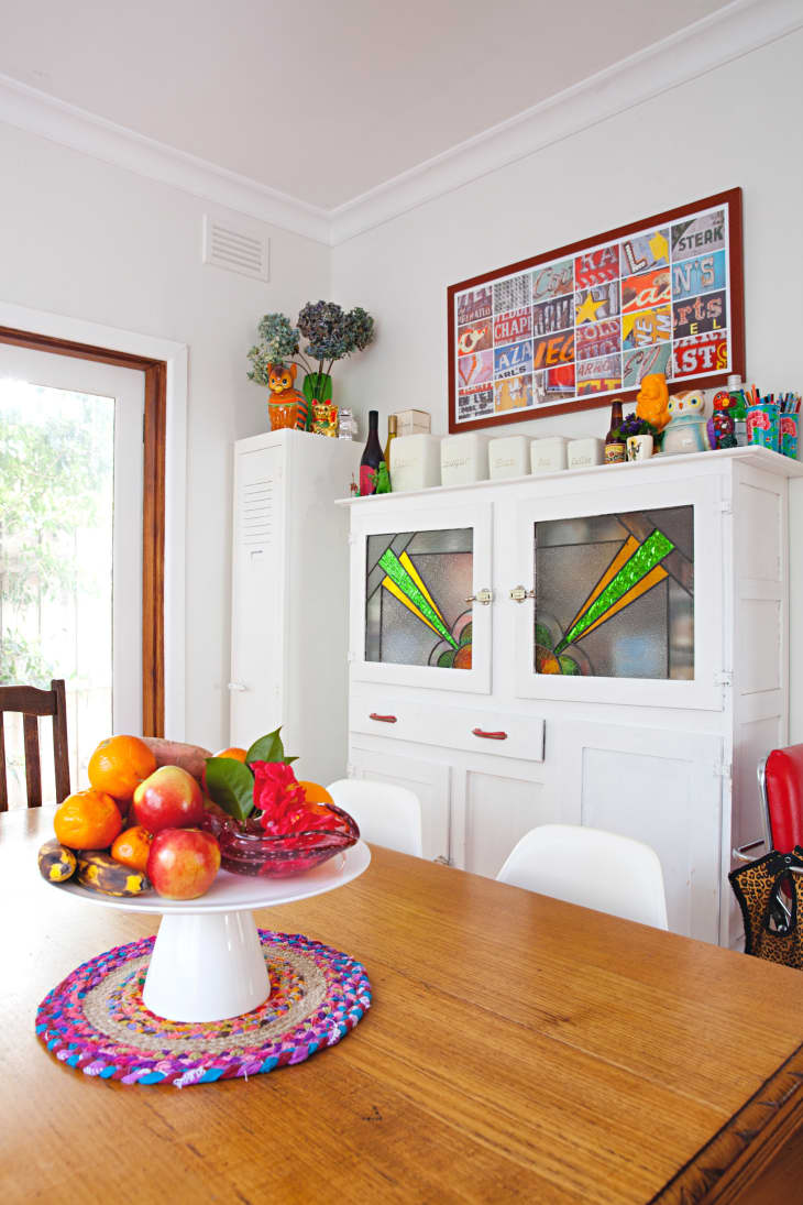 Dining room with wooden table, fruit centerpiece, white cabinet with stained glass, and colorful wall art.