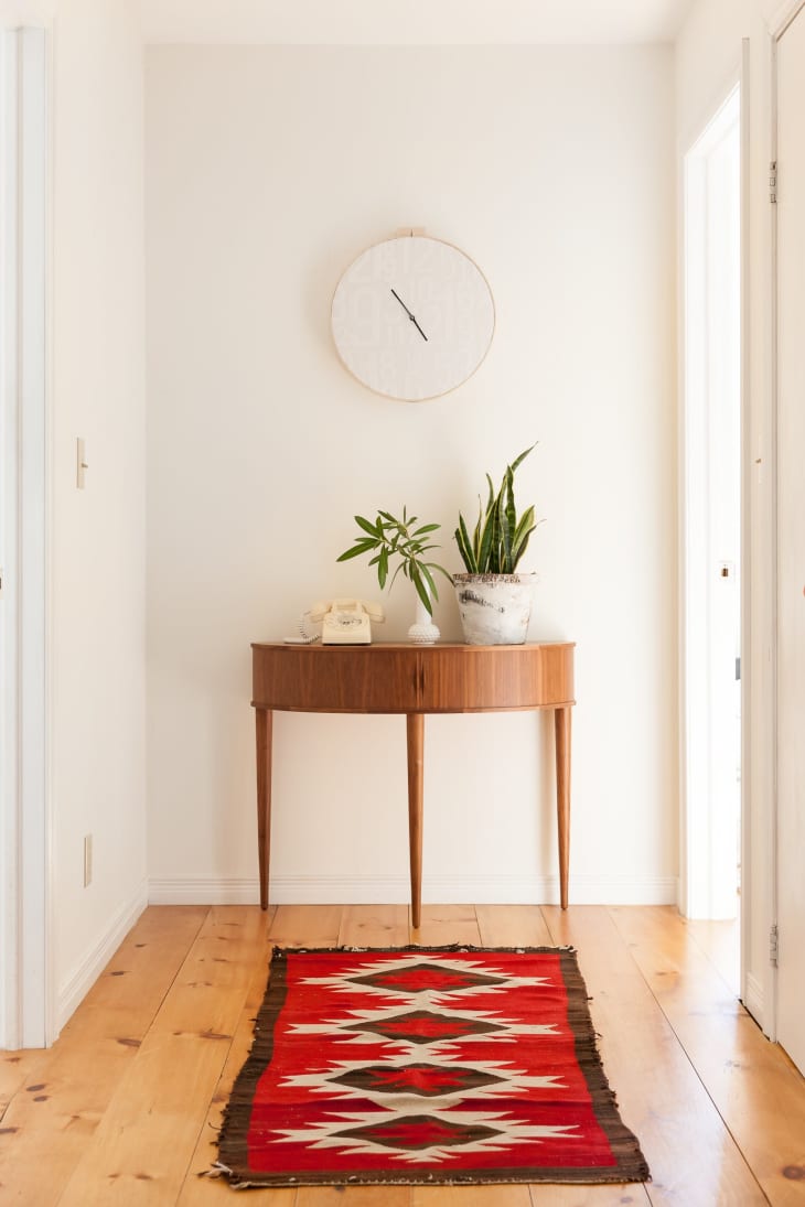 Hallway with wooden console table, potted plants, vintage phone, round wall clock, and red geometric-patterned rug.