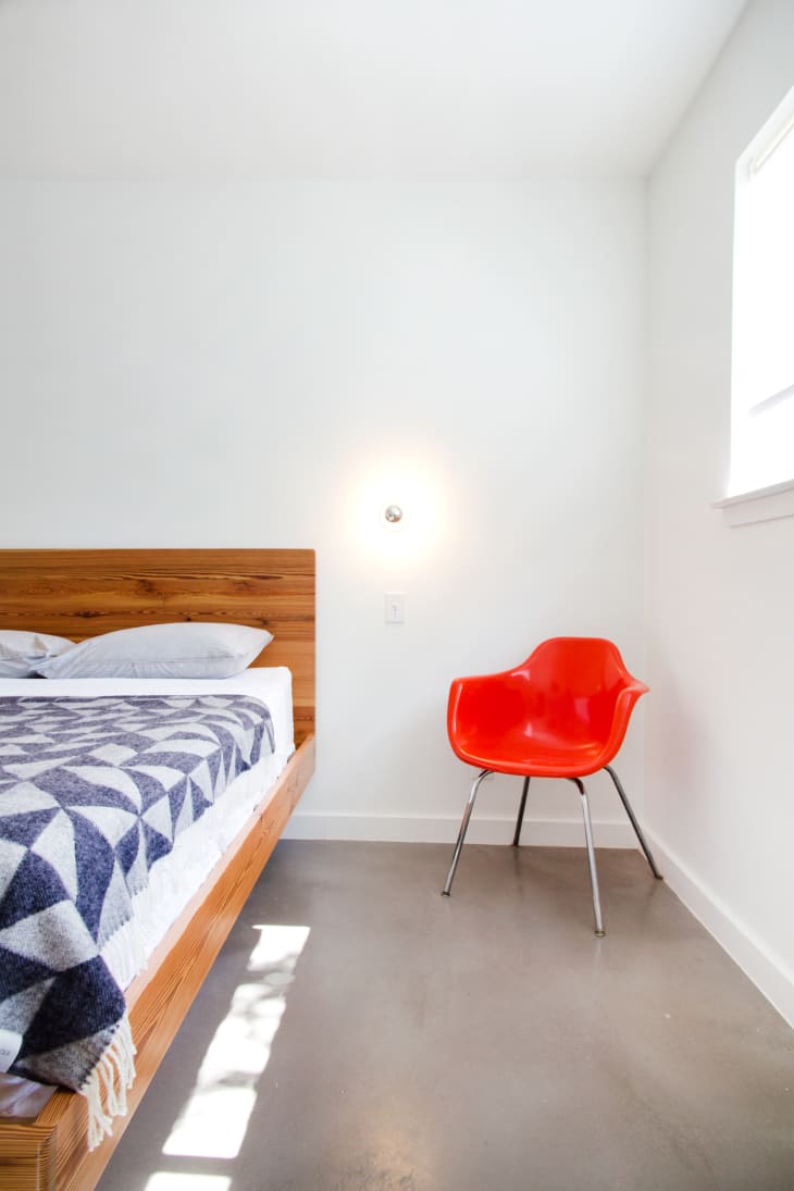 Minimalist bedroom with a wooden bed, geometric blanket, and a red chair under a wall light.
