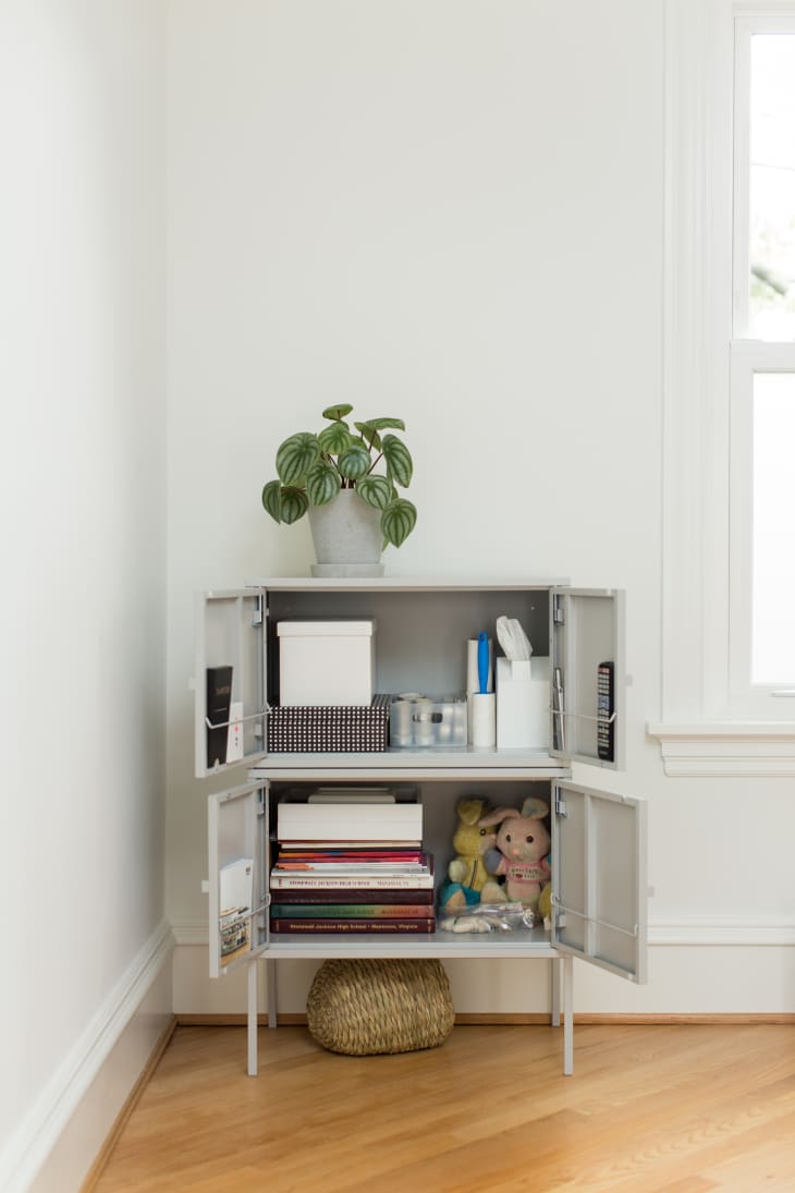 Gray cabinet with open doors, displaying books, toys, and office supplies, topped with a potted plant.