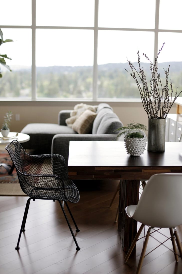 Modern living room with a gray sofa, black wire chair, wooden table, and decorative plants near large windows.