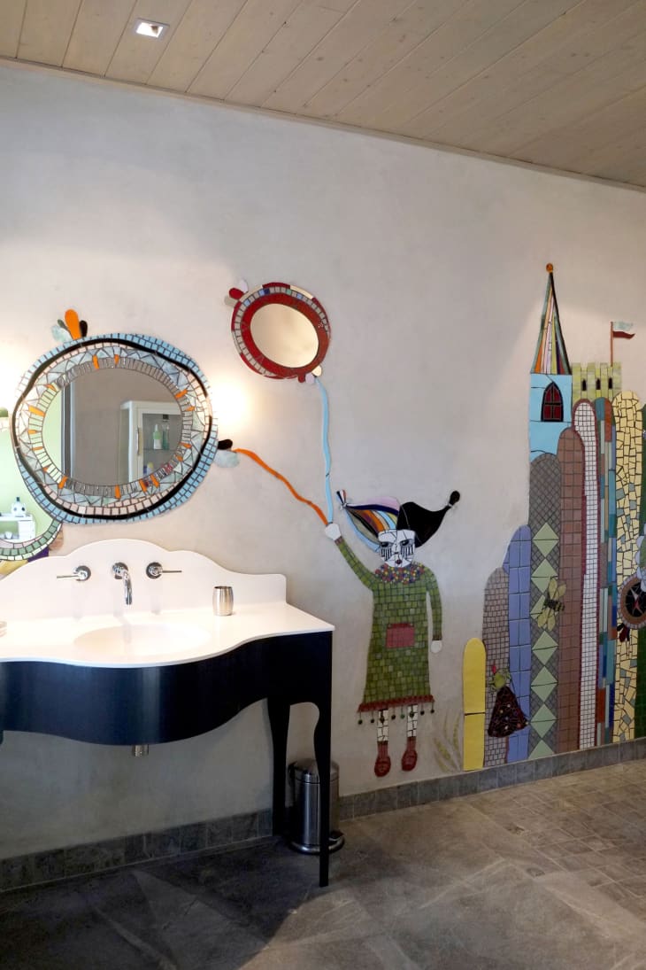 Bathroom with mosaic wall art, featuring a whimsical figure holding a balloon, and a curved sink with a round mirror.