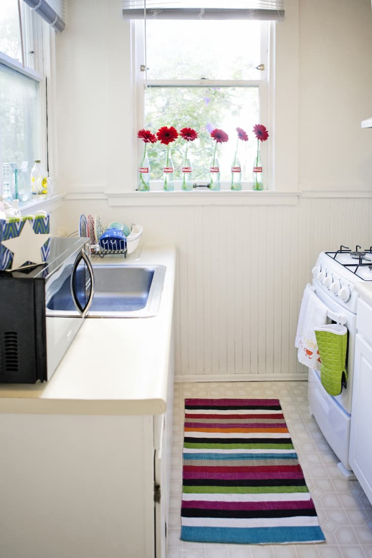 Small kitchen with striped rug, red flowers in bottles on windowsill, blue sink, and white stove.
